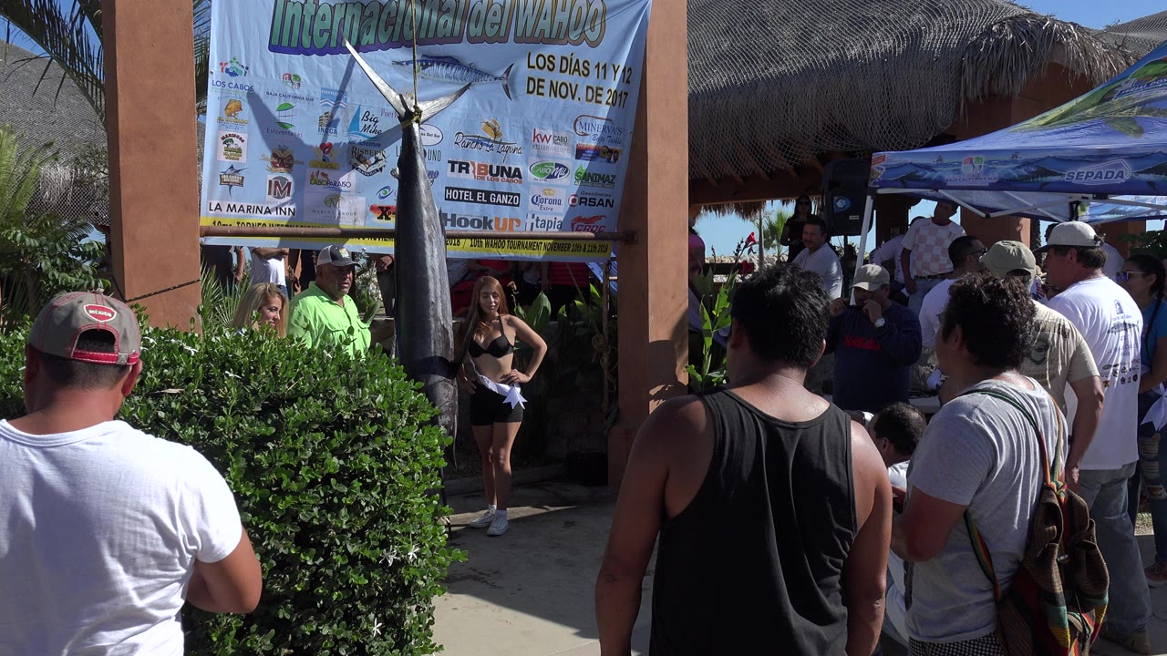 At 0:01, the Playita ceremony remains framed from behind the crowd, with a woman in black and sash near the banner and several adults gathered under the canopy.