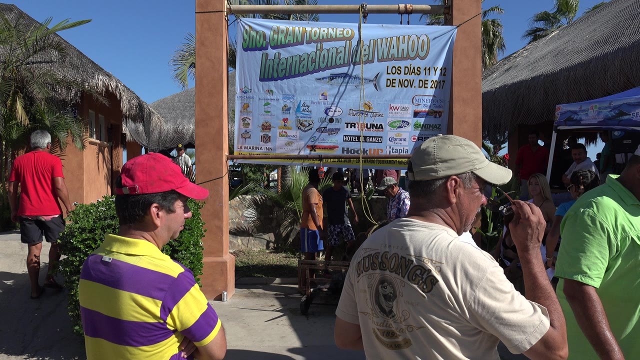 At 0:00 in Playita, Baja California Sur, people stand and walk beneath a large Spanish-language tournament banner beside thatched-roof structures.