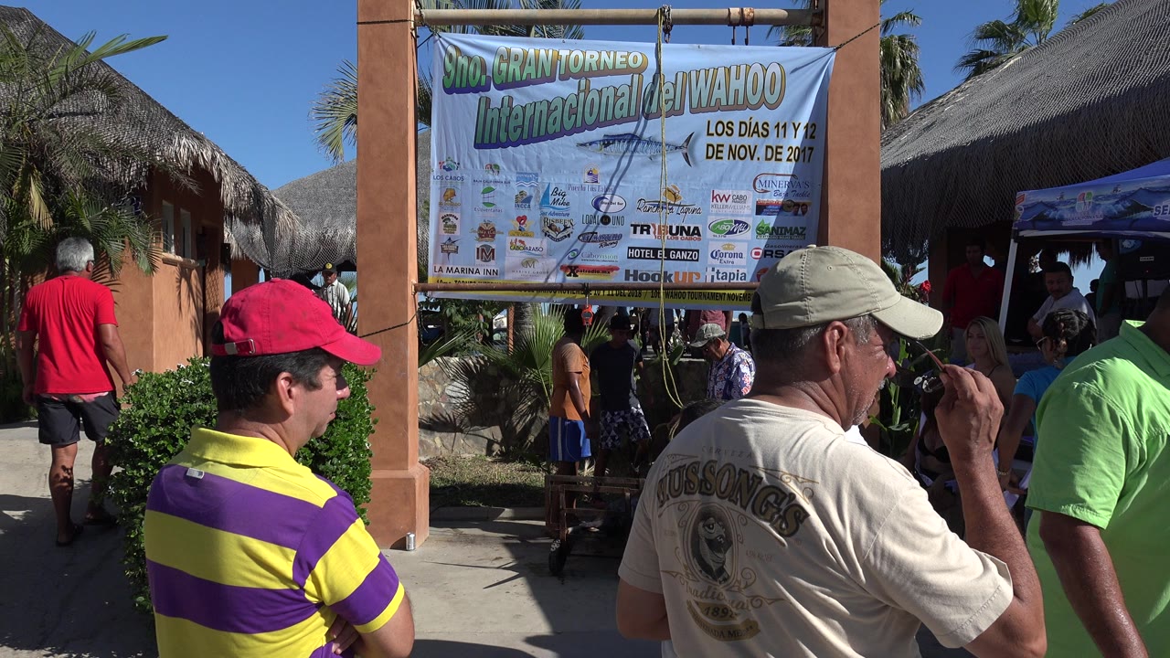 At 0:01 in Playita, Baja California Sur, a man in a light cap raises a hand near his face while other people move around the bannered entrance.