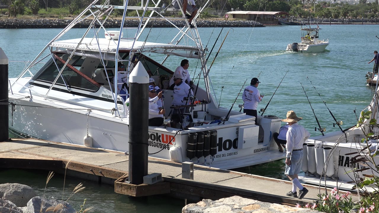 At 0:01 in Playita, Baja California Sur, the man continues along the dock as adults remain on the fishing boat and a small boat moves across the water in the background.
