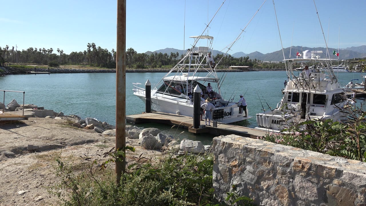 At 0:01 in Playita, Baja California Sur, the boats remain moored beside the dock while people cluster near the stern area, with the foreground post and stone wall framing the view.
