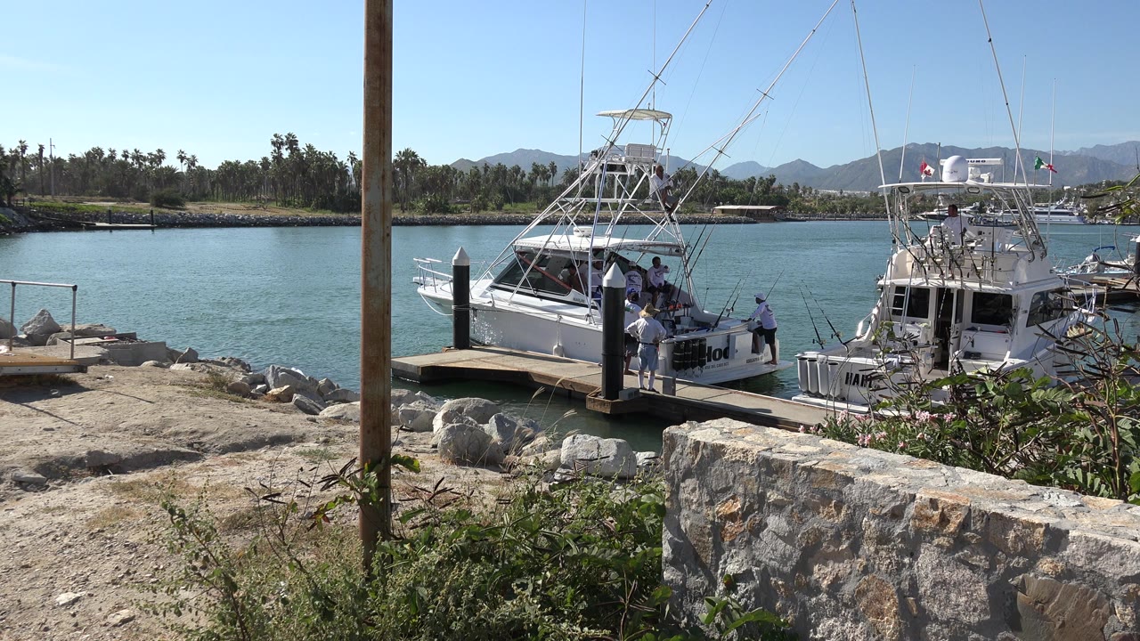Two white fishing boats docked beside blue-green water with people on board and on the pier.