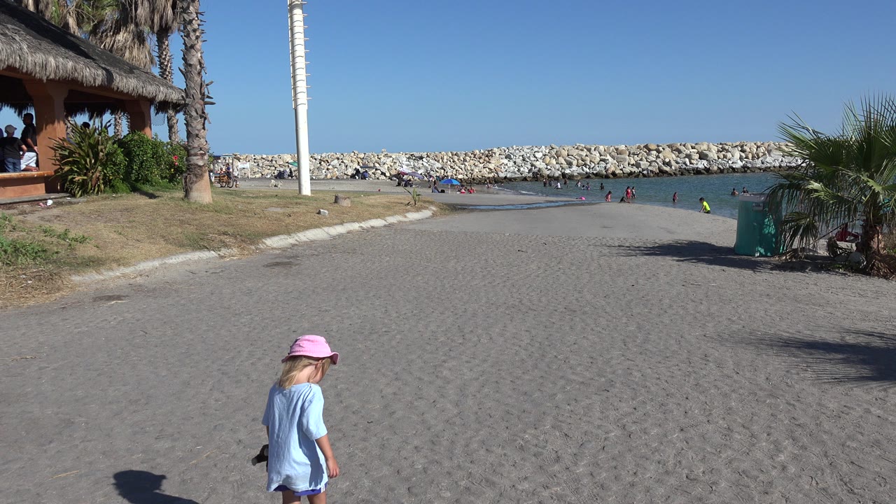 At 0:01 in Playita, the girl continues along the sand near the lower center of frame while the rock breakwater, shallow water, beach hut, palms, and scattered people remain visible behind her.