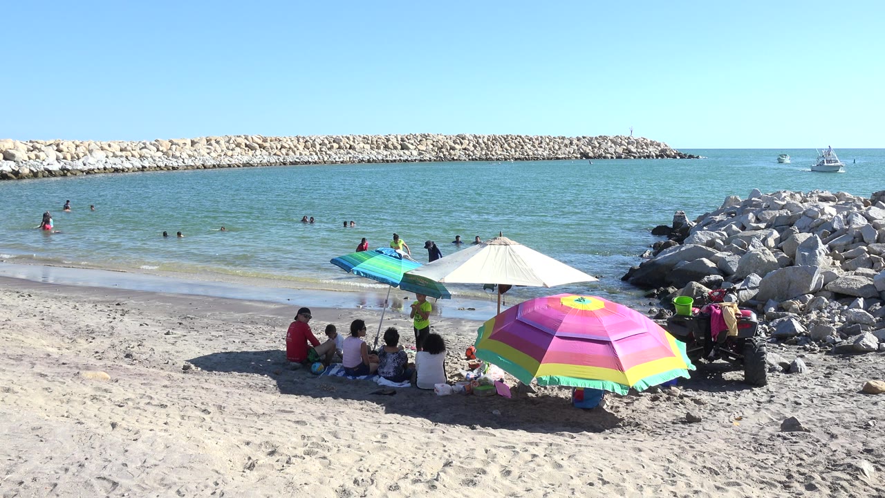 Playita beach at one second with the foreground umbrellas partly shading seated beachgoers while swimmers remain in the shallow blue-green water near the breakwater.