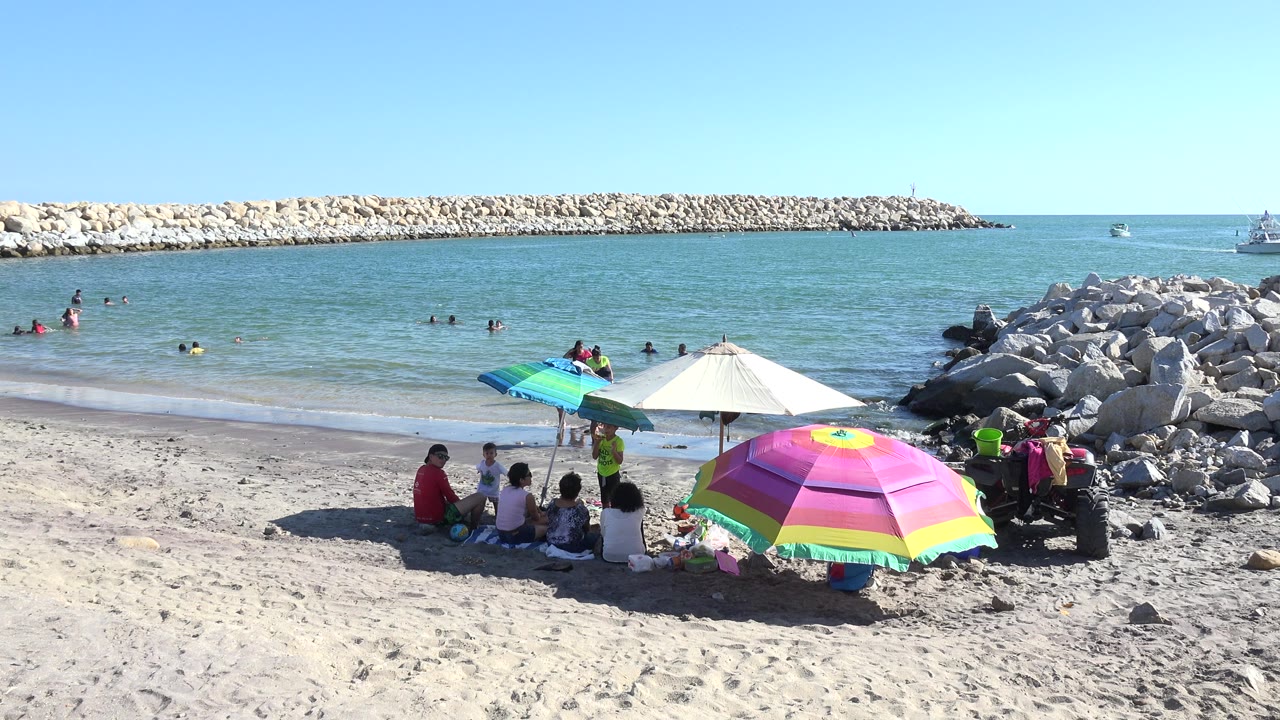 Beachgoers under colorful umbrellas beside blue water and rock jetties.