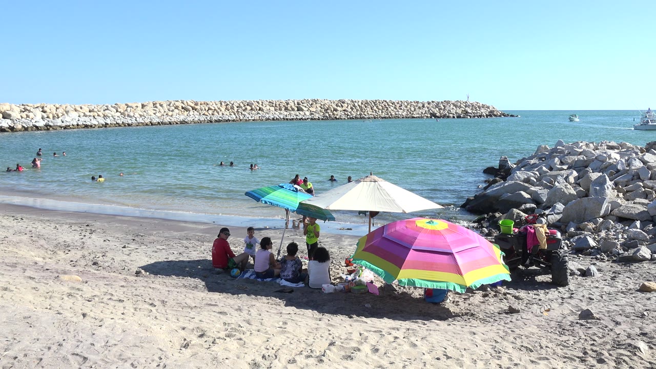 Playita beach scene with the rainbow umbrella prominent in the foreground, a white umbrella near the rocks, and scattered people wading in the blue water.