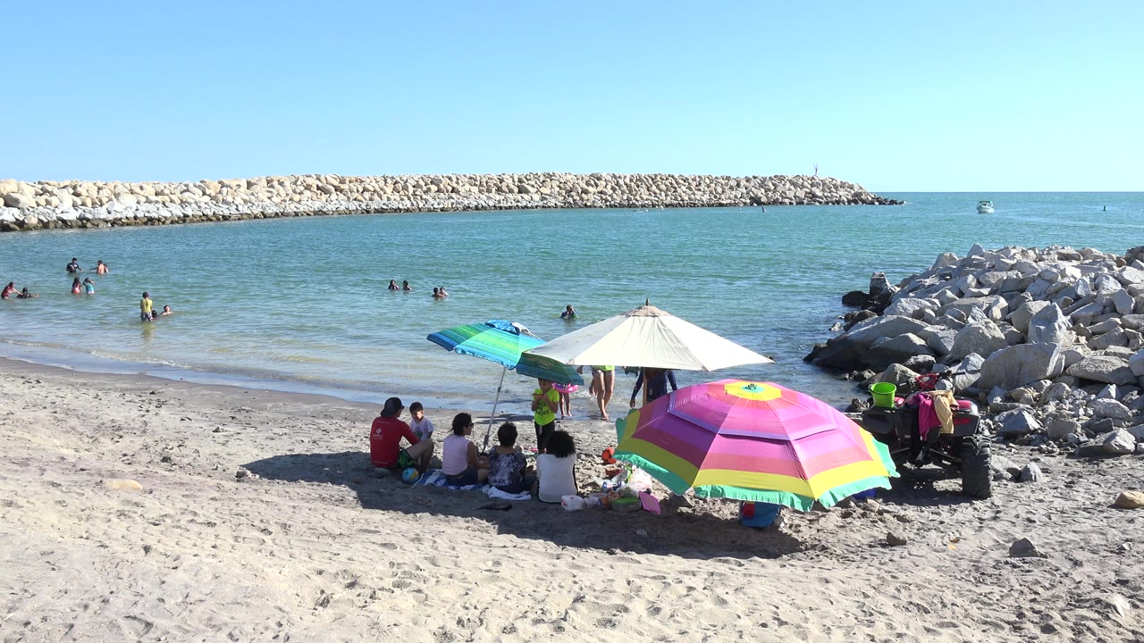 Family group under colorful beach umbrellas beside swimmers and a rock breakwater.