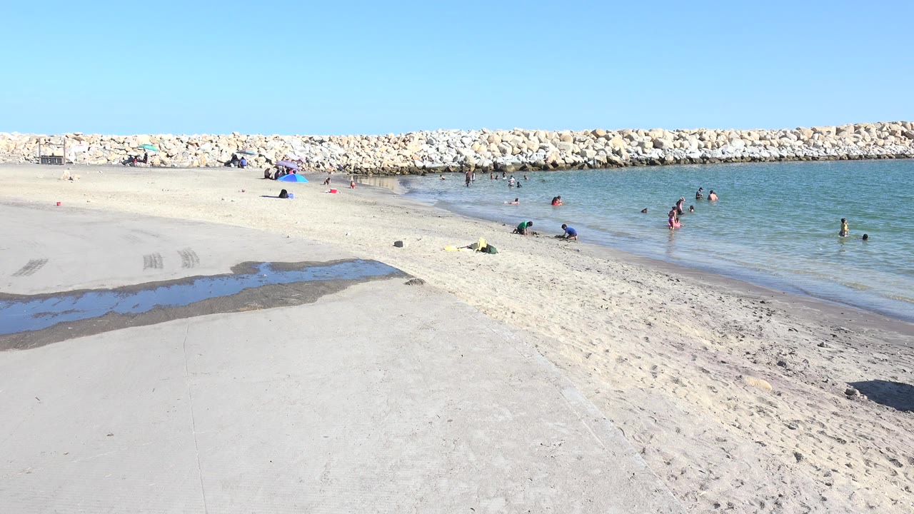 Wide sandy beach with swimmers near a rock breakwater