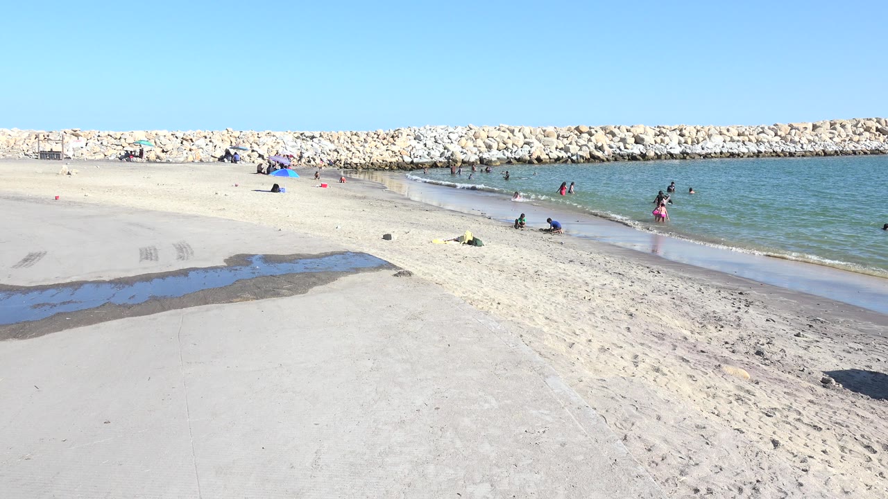 Opening frame of a Playita, Baja California Sur beach with white sand, a shallow wet channel, calm blue-green water, distant beachgoers, and a pale rock breakwater.