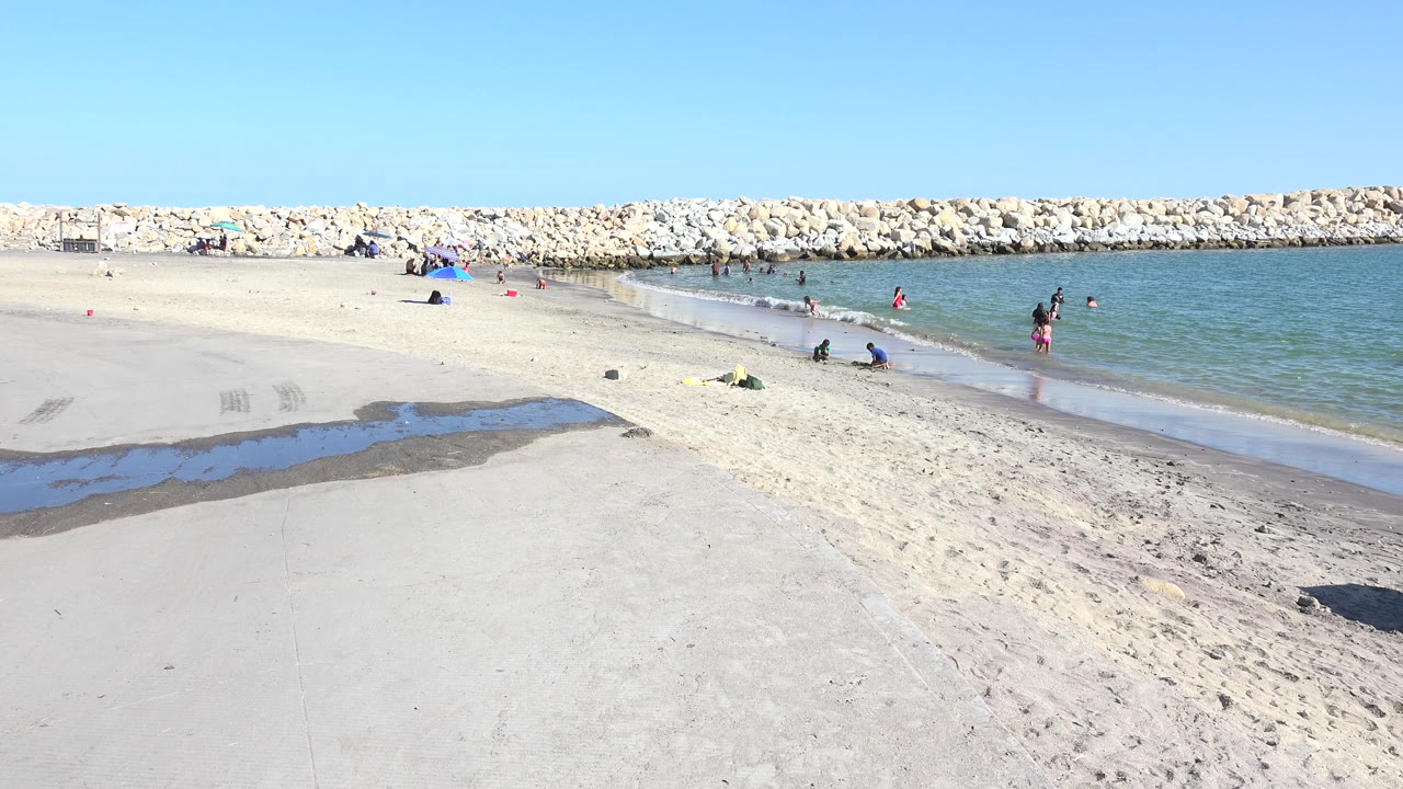 White beach, calm water, and a long rock breakwater under clear sky.