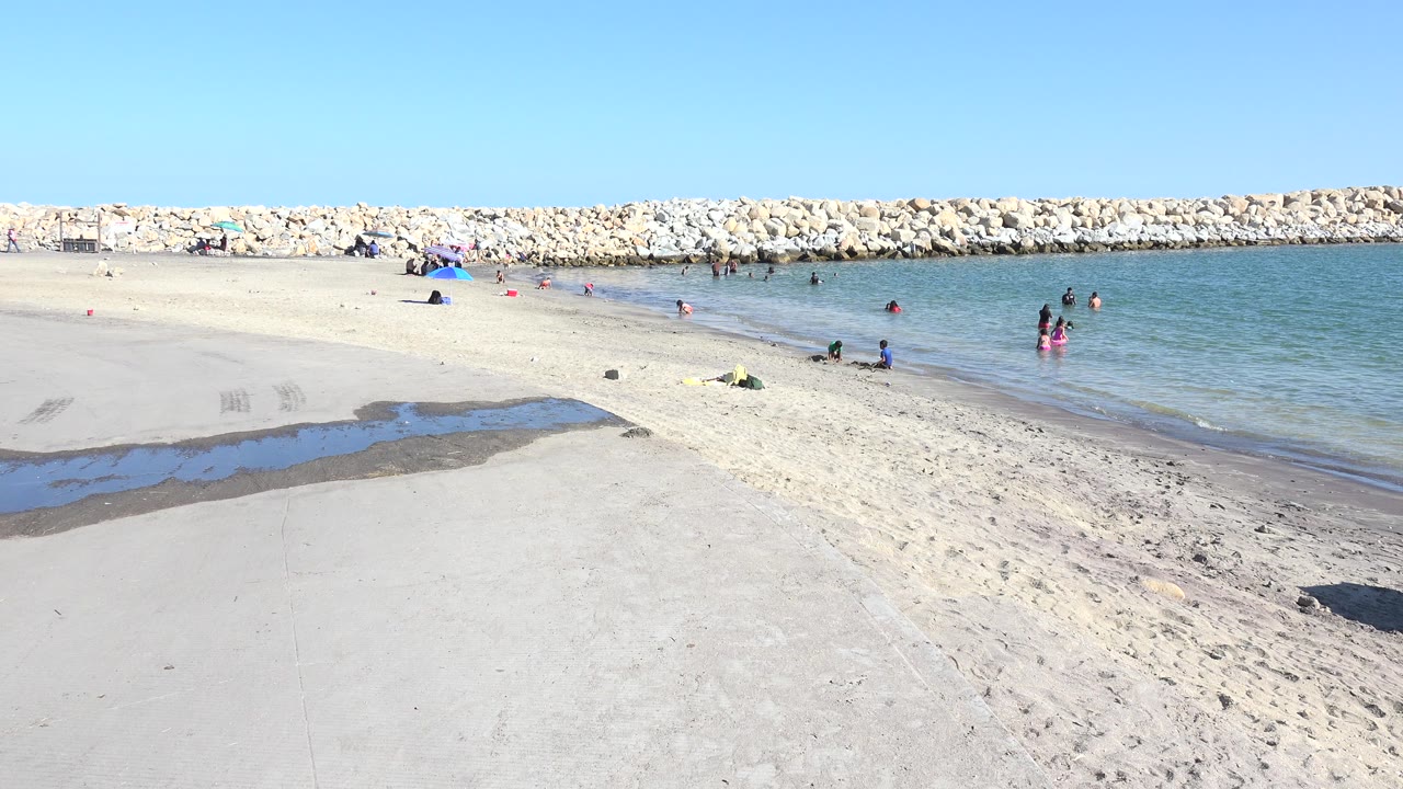Wide sandy beach with people in calm water near a rock breakwater.