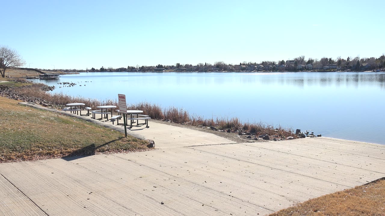 Opening frame at Hidden Lake, Colorado showing a pale lakeside ramp, picnic tables near reeds, calm blue water, a distant tree line, and no visible people.