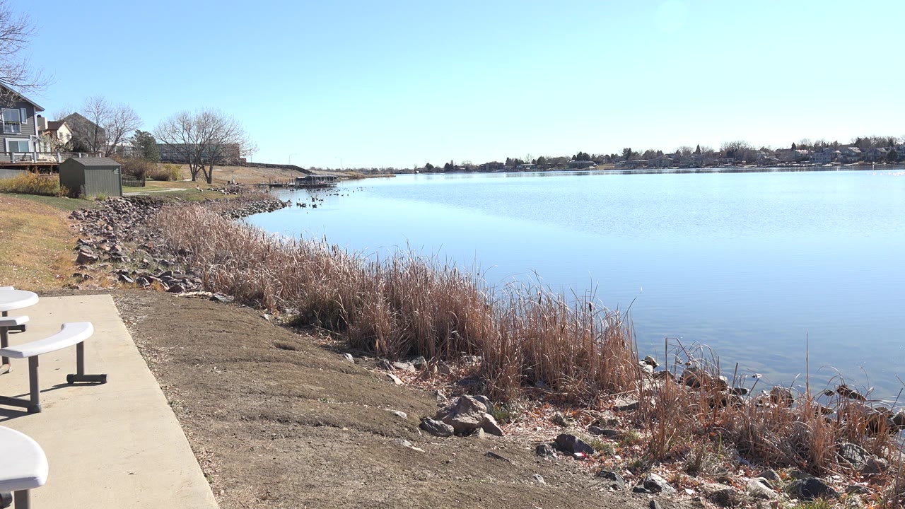 Opening frame of Hidden Lake, Colorado with a concrete path and partial picnic tables at left, dry shoreline grass and rocks in front, calm blue water to the right, distant homes, and no visible people.