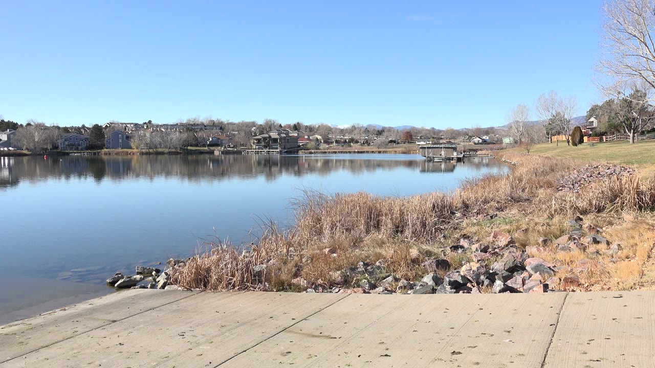 Opening view of Hidden Lake, Colorado from a concrete boat ramp, with dry reeds and rocks at the shore, a small dock across the water, distant houses, leafless trees, and no visible people.