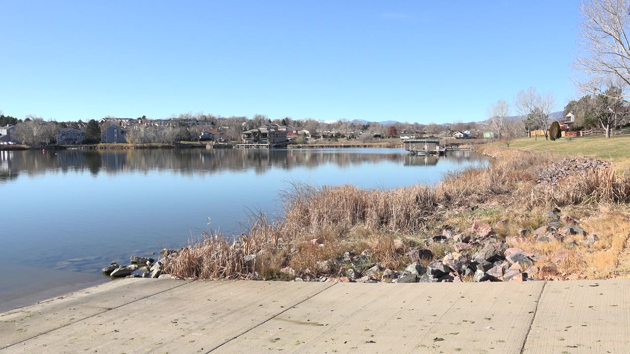 Still lake beside a concrete ramp, reeds, rocks, distant houses, and clear blue sky.