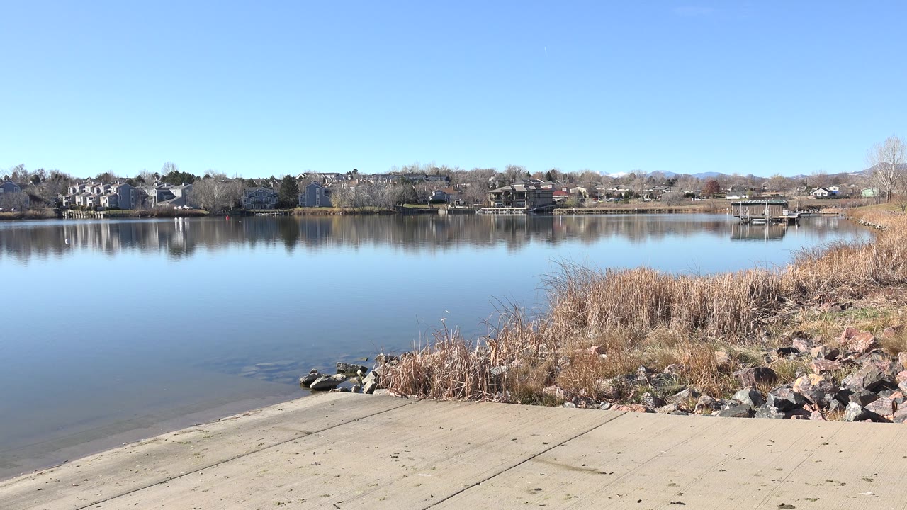 At 0:01, Hidden Lake, Colorado remains framed from the shoreline, with smooth reflective water, reeds and rocks on the right edge, a small dock across the lake, distant houses, and no visible people.
