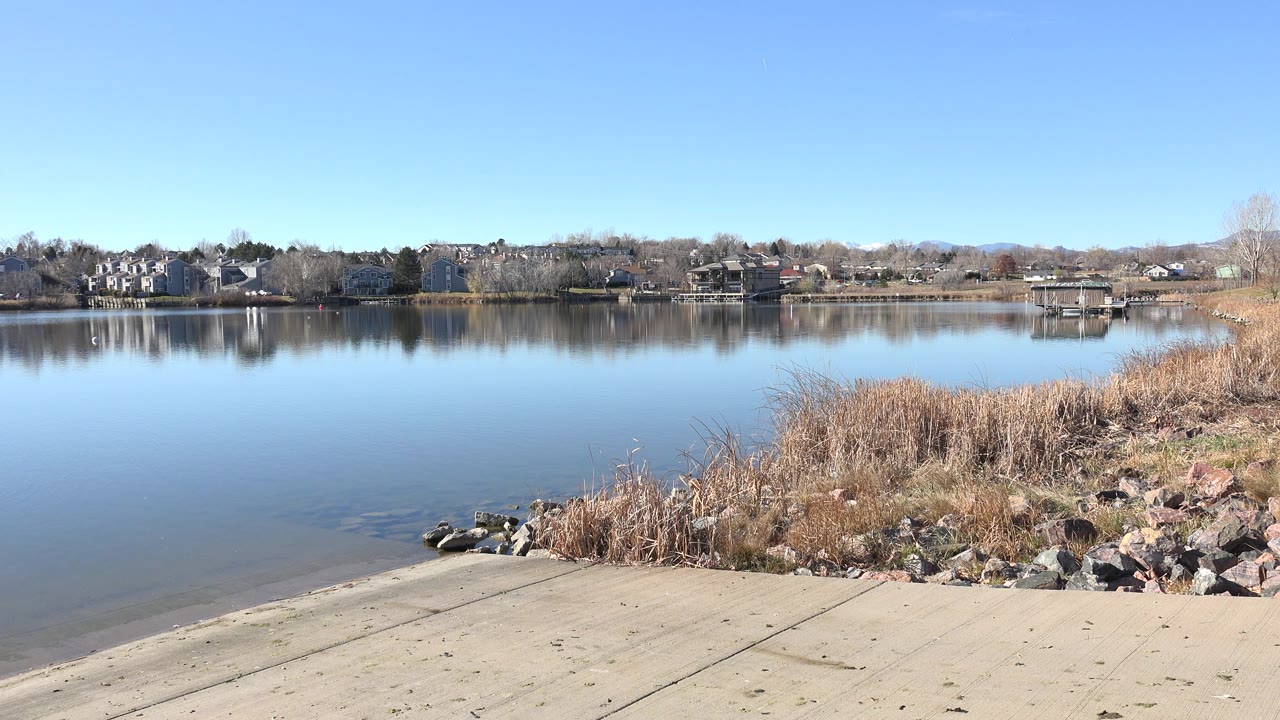 Lakeside access ramp with reeds, rocks, still water, and distant houses.