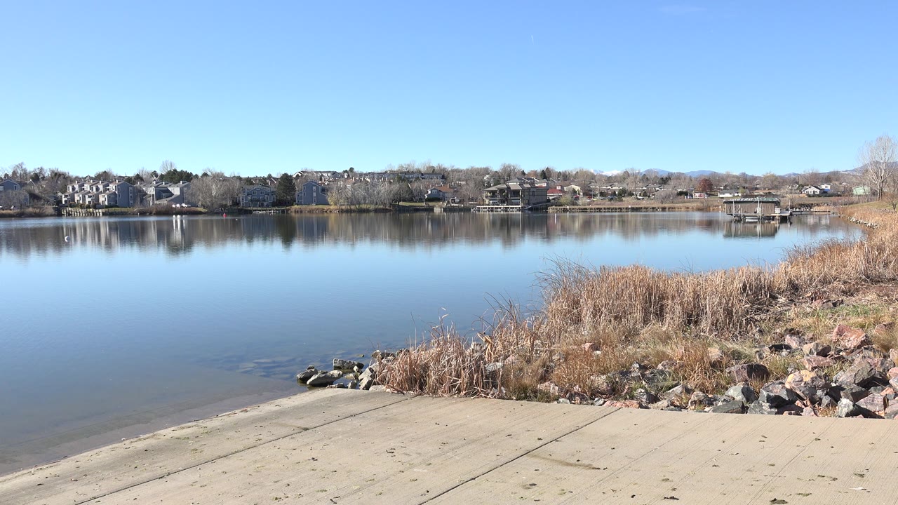 Opening view of a concrete boat ramp at Hidden Lake, Colorado with still reflective water, shoreline reeds, rocks, distant homes, bare trees, and no visible people.