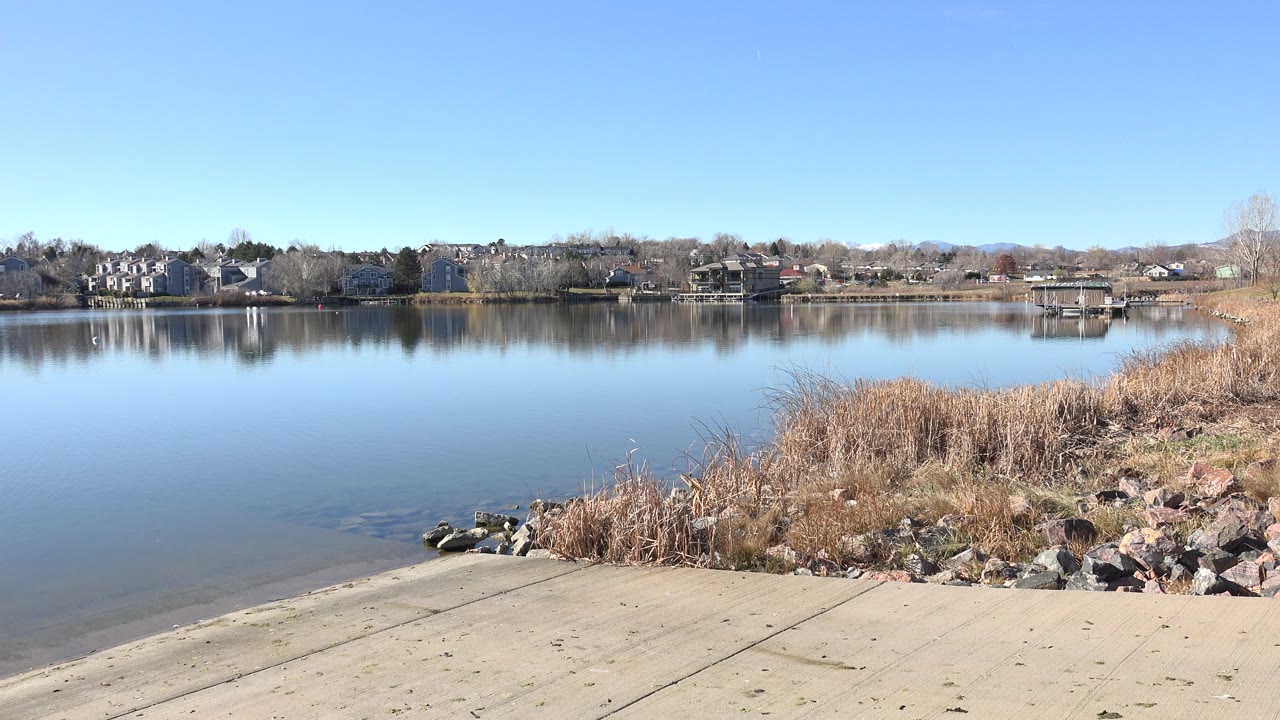 Second frame holds on the Hidden Lake boat ramp with the waterline at left, tan reeds and rocks at right, distant residential buildings, and no visible people.