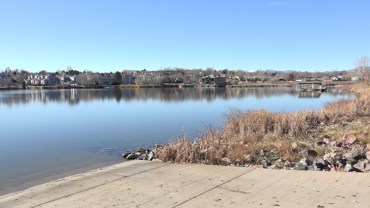 Concrete boat ramp beside calm lake water with reeds and distant homes.