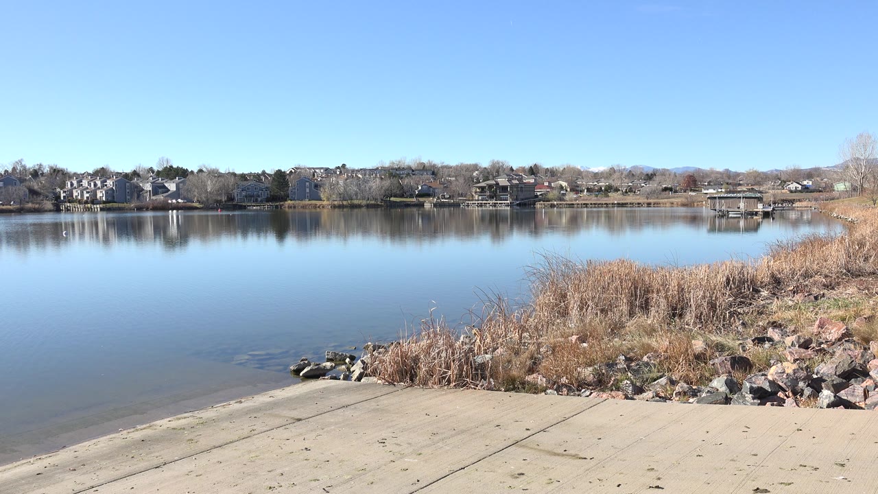 Hidden Lake, Colorado frame with the ramp in the lower foreground, smooth blue water across the center, a reed bed at right, far docks and homes, and no visible people.