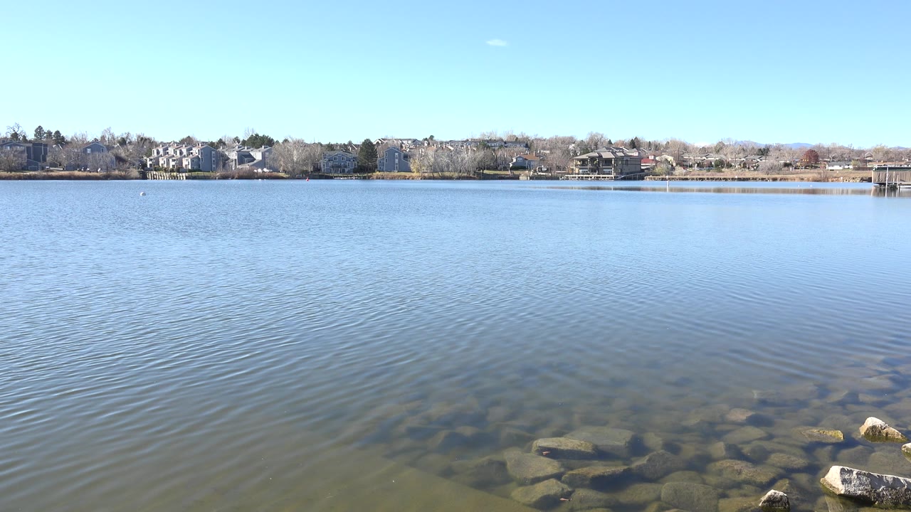 At 0:01, the rippled lake surface spreads across the frame while foreground stones remain visible under shallow water and homes line the far shore, with no visible people.