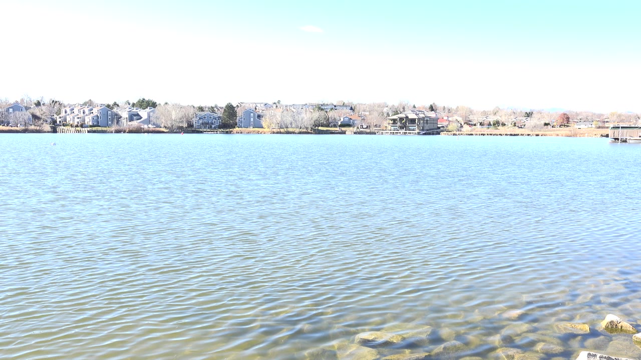 Bright lake water with rocks in the foreground and houses across the shore.