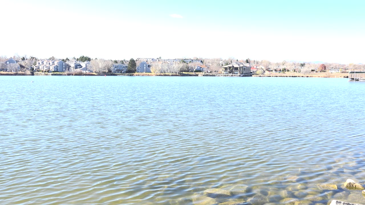 Second frame of Hidden Lake, Colorado showing the same broad water view with small ripples across the lake, a pale sky, far shoreline homes, trees, and no visible people.