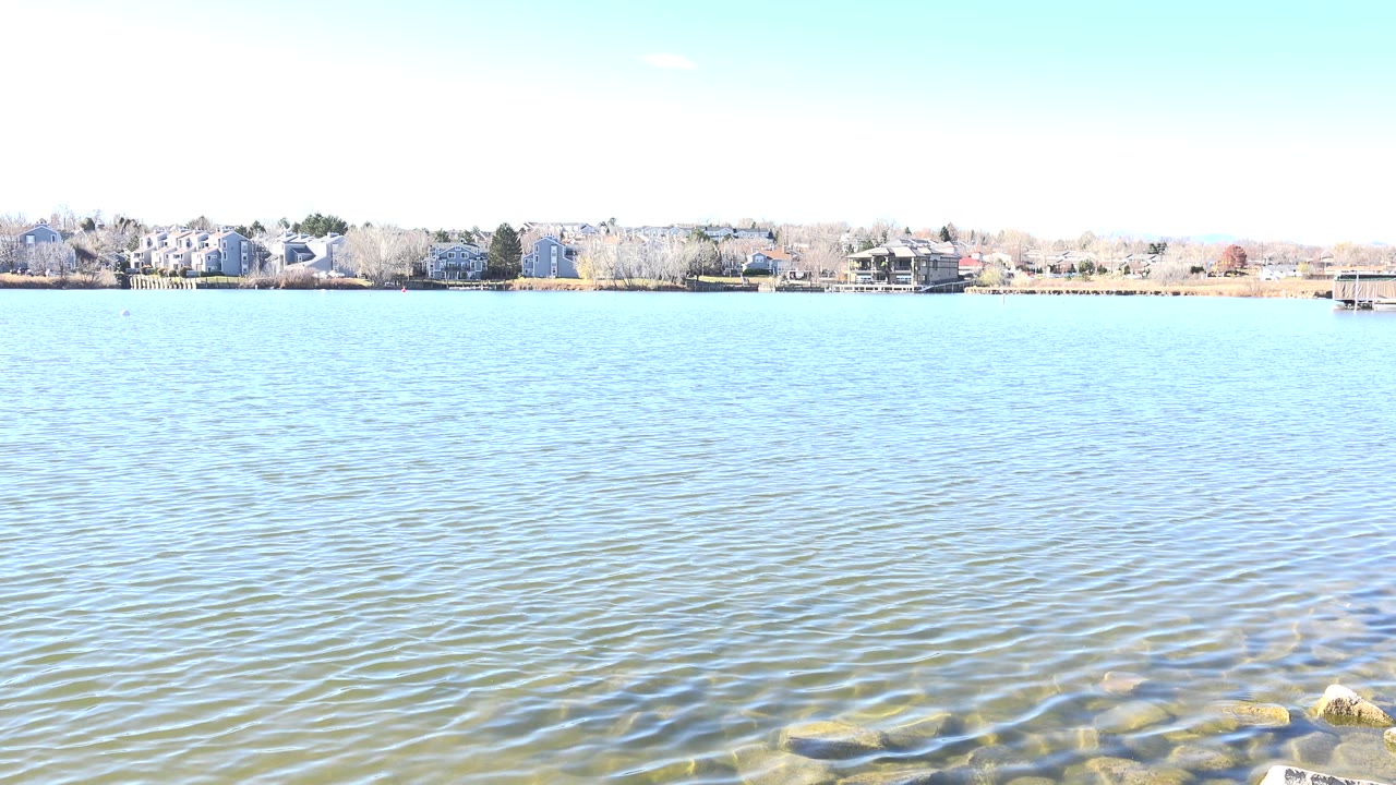 Opening frame of Hidden Lake, Colorado with clear rippled water, submerged shoreline rocks, distant homes and trees, and no visible people.