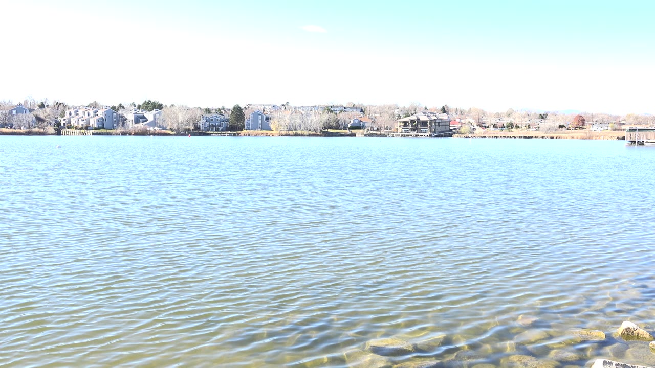 Second frame on Hidden Lake, Colorado showing a wide band of pale blue water, foreground rocks, distant shoreline buildings, and no visible people.