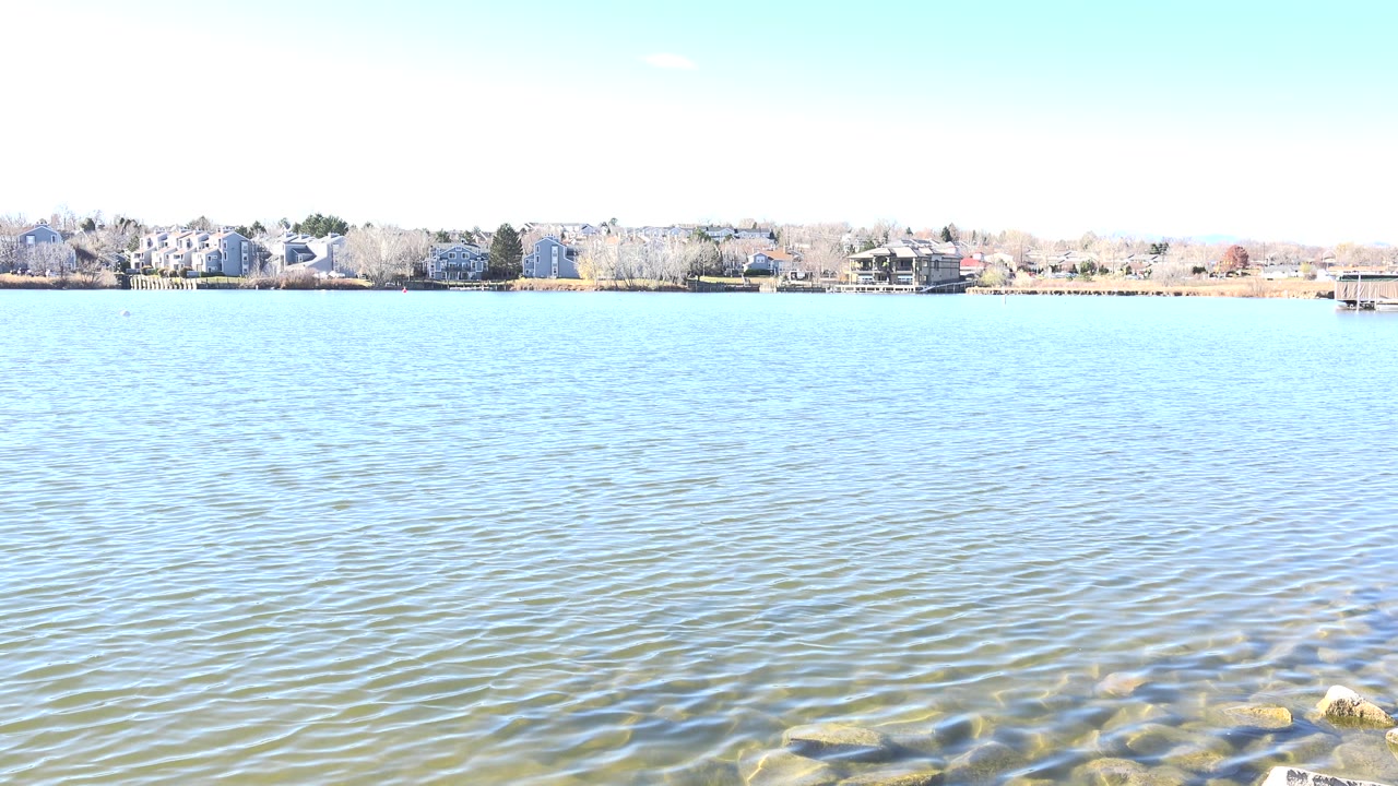 Wide lake view with shallow rocks at lower right and homes along the far bank.