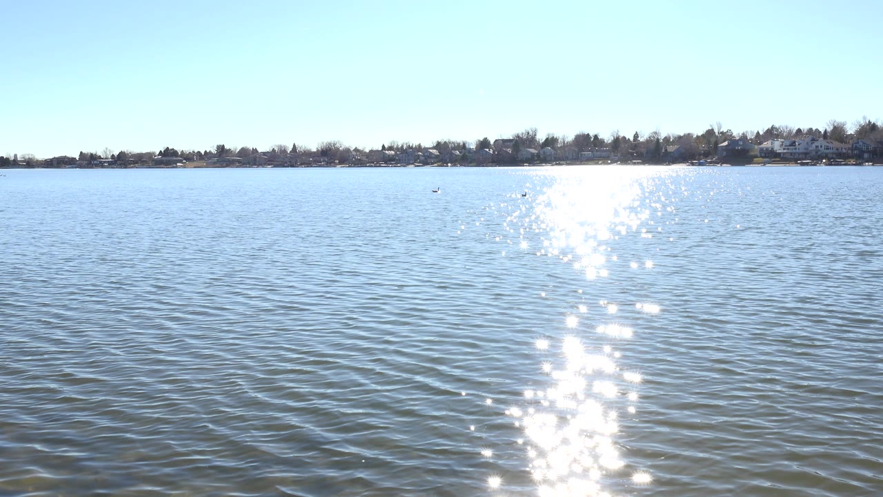 Opening frame of Hidden Lake Colorado with rippled blue water, a bright column of sun reflection near center, a low tree-lined shoreline, pale sky, and no visible people.