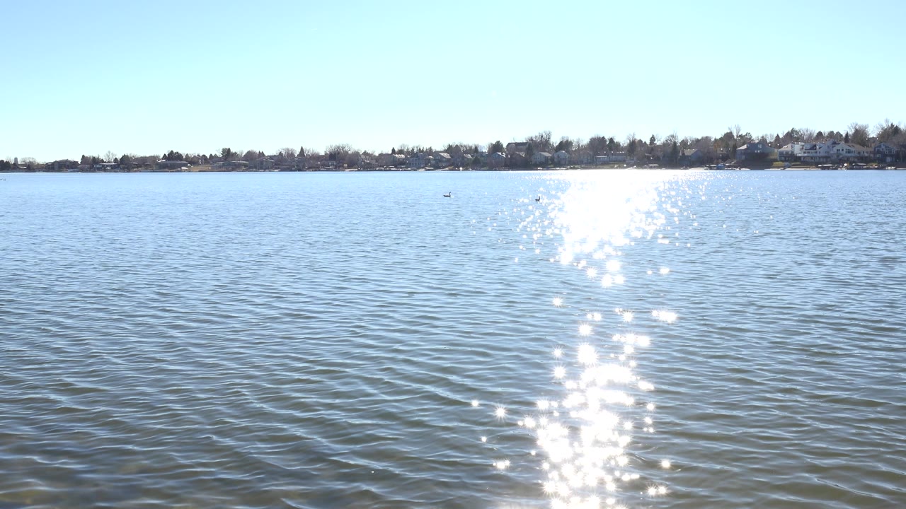 Hidden Lake Colorado water fills the frame with a strong white sun sparkle running down the center and a thin distant shoreline behind it, with no visible people.