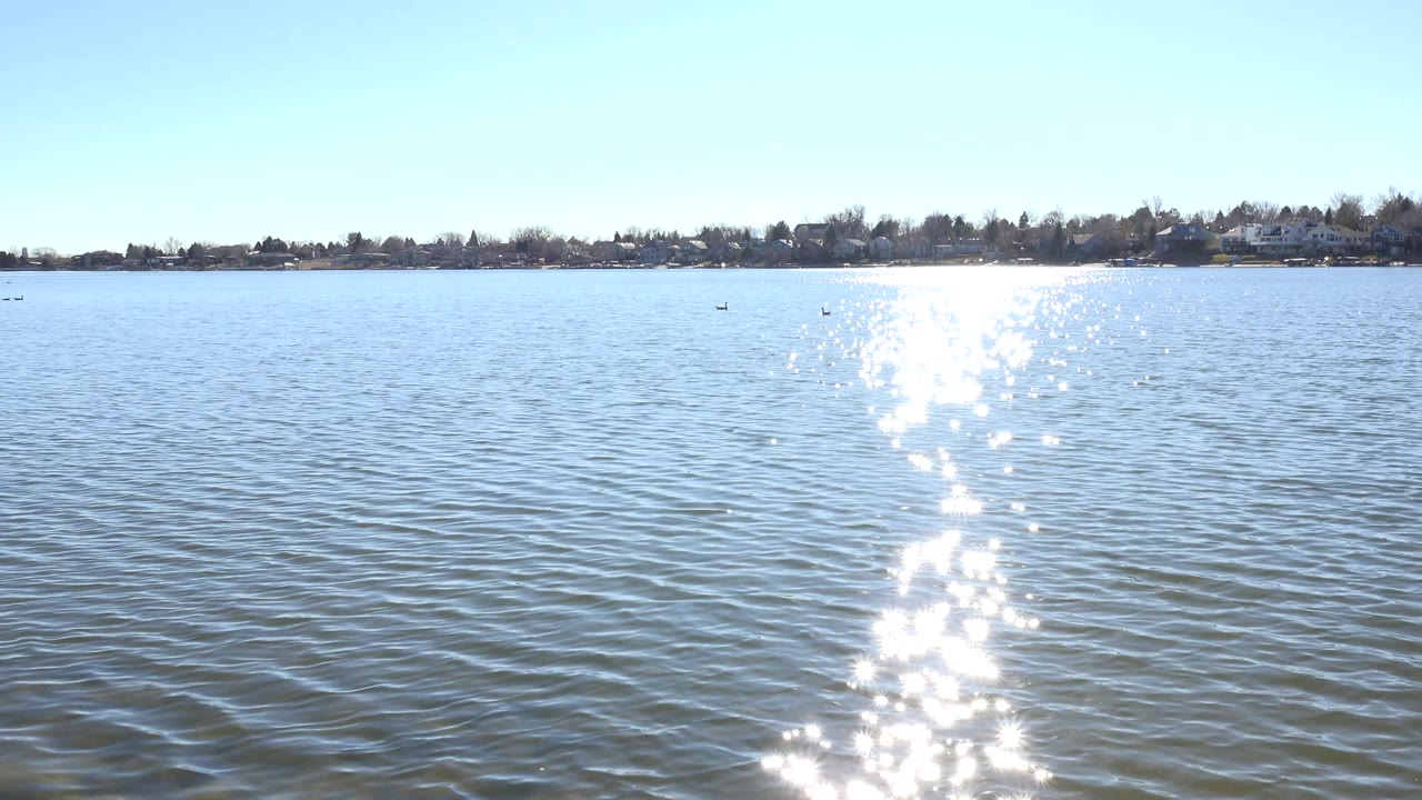 Opening frame of Hidden Lake, Colorado with rippled water in front, a bright vertical sun glint near center, distant shoreline buildings, and no visible people.