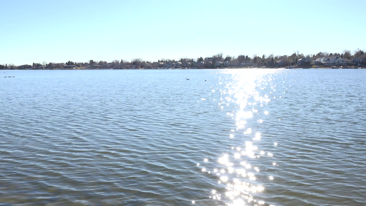 Hidden Lake, Colorado at 0:01 shows the sun reflection slightly broken by ripples, open blue water across the foreground, a narrow far shoreline, and no visible people.