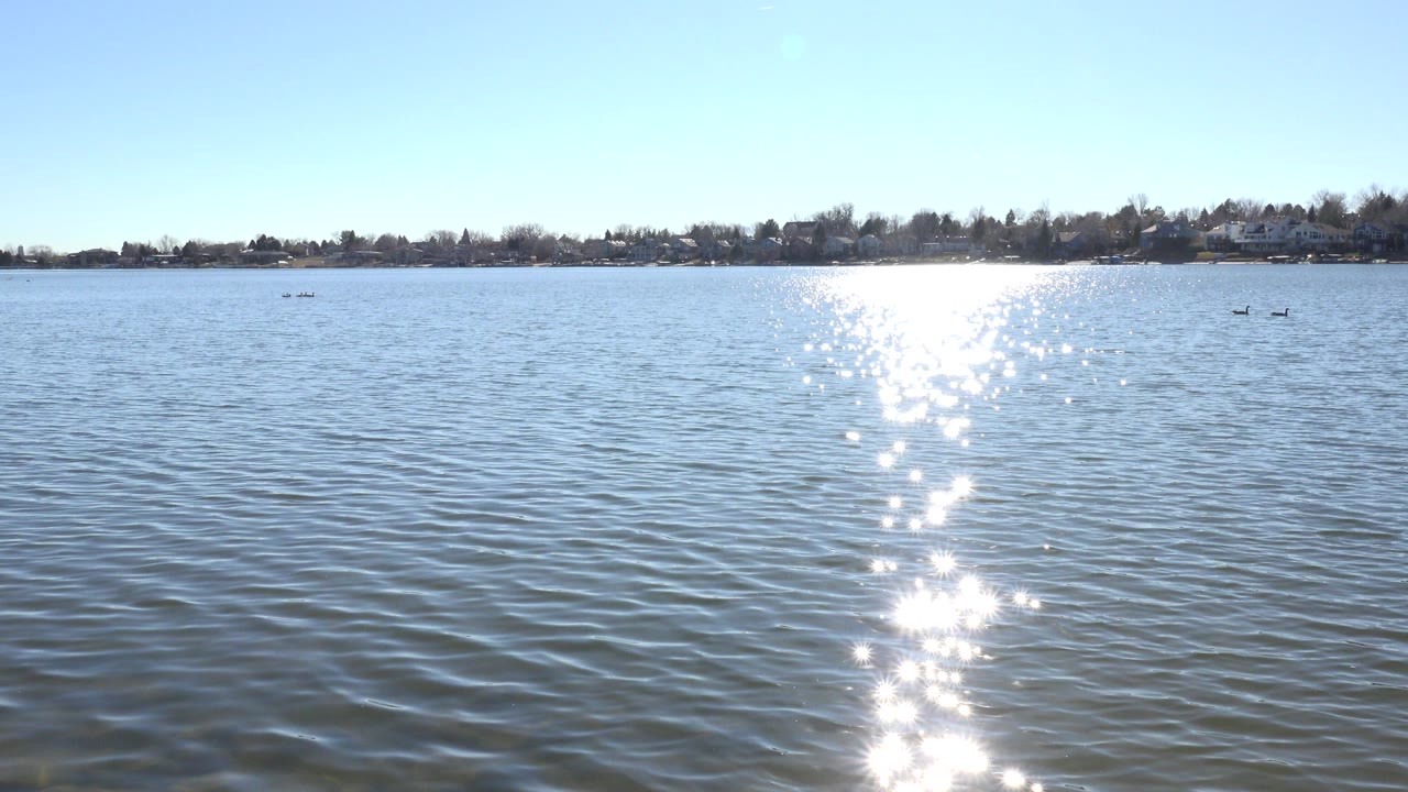 Bright sun reflection across rippled lake water with a distant tree-lined shore.