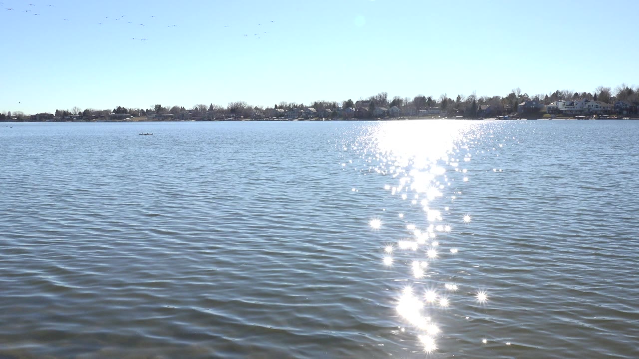 At 0:00, Hidden Lake in Colorado fills the frame with rippled blue water, a bright vertical sun reflection, distant shoreline trees and homes, and no visible people.