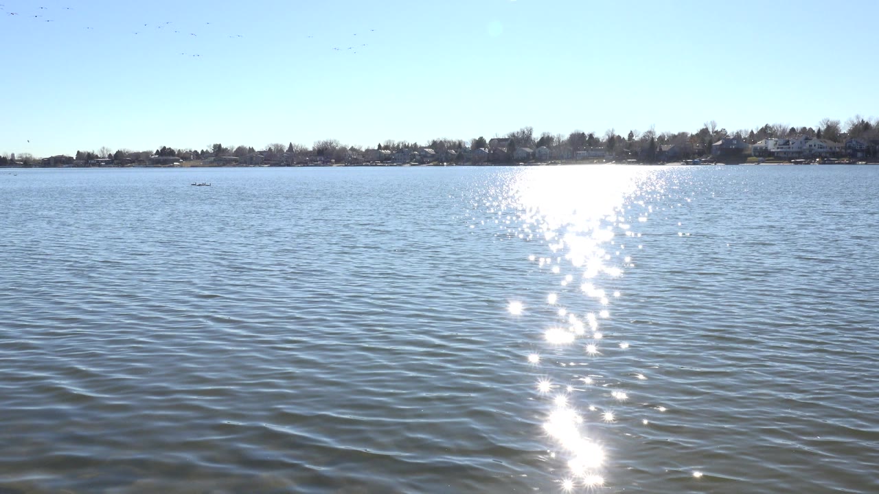 At 0:01, the Hidden Lake view holds on blue ripples, a centered band of sun sparkle, a low tree-lined horizon, distant homes, and no visible people.