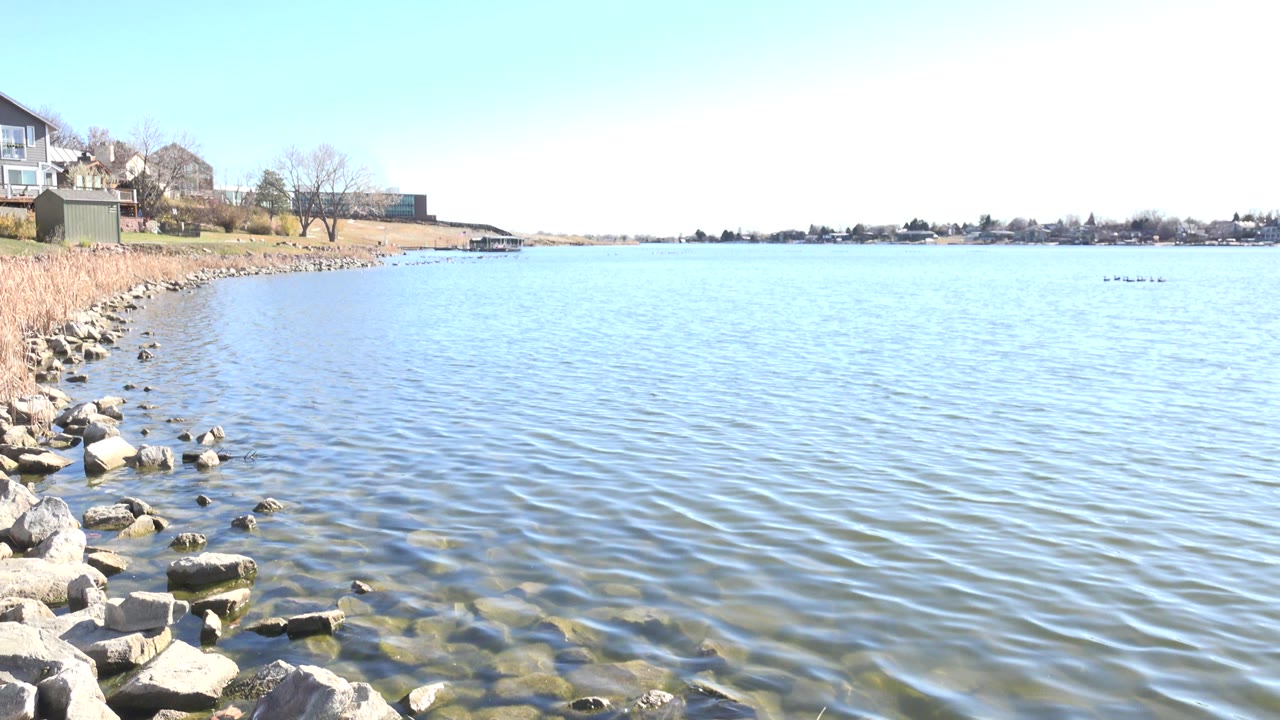 Opening frame at Hidden Lake, Colorado showing rippling blue water, submerged foreground rocks, a rocky reed-lined shore at left, distant houses and trees, and no visible people.