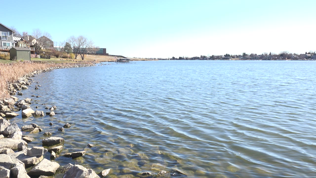 Opening frame of Hidden Lake, Colorado with shoreline rocks in the foreground, rippled blue water across the frame, houses on the left bank, distant trees on the horizon, and no visible people.