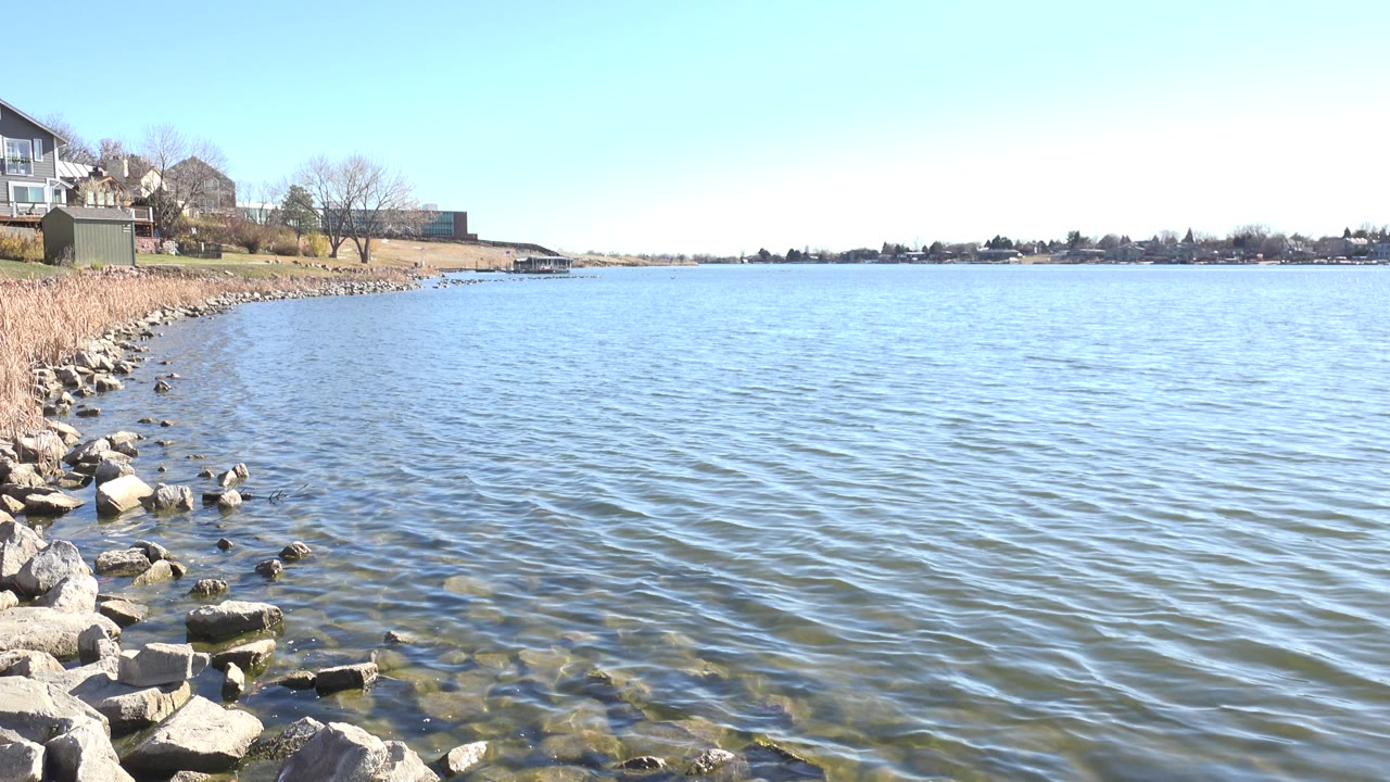 Hidden Lake, Colorado shoreline at one second, showing small ripples across blue water, rocks in the lower left, reeds on the bank, distant houses, and no visible people.