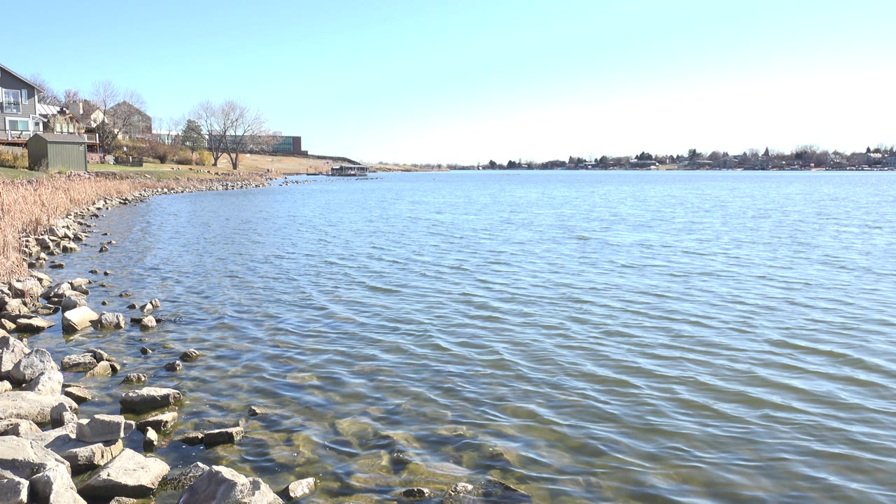 Hidden Lake Colorado shoreline at 0:01 with shallow clear water over submerged rocks, pale shoreline stones at left, reeds behind them, and no visible people.
