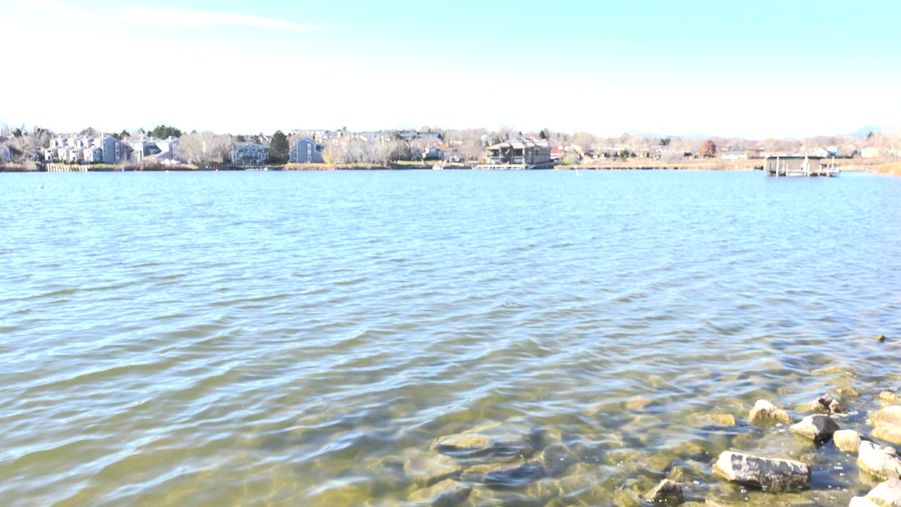 Hidden Lake, Colorado at 0:01 shows small wind ripples across the water, shoreline stones at the lower right, far houses, trees, docks, and no visible people.