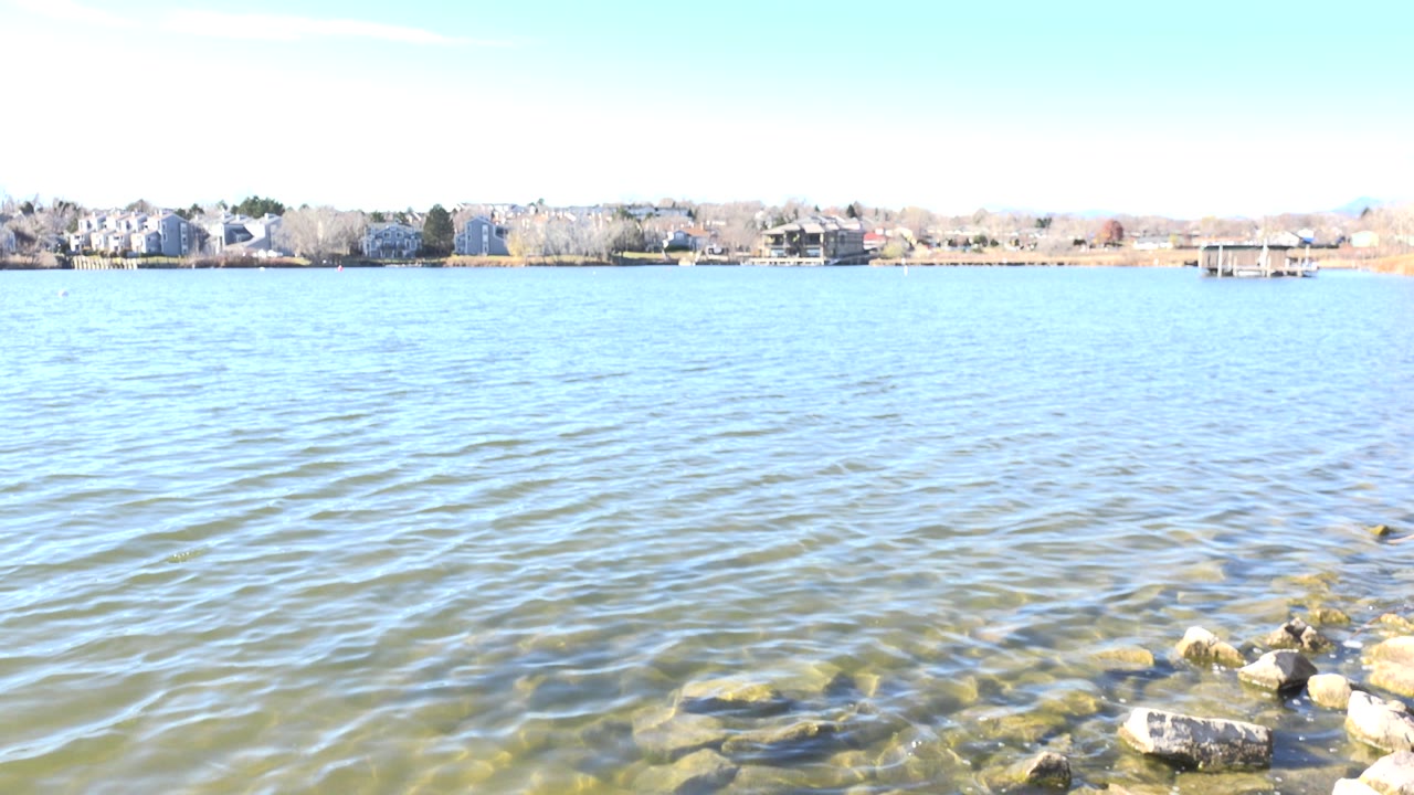 Opening frame of Hidden Lake, Colorado showing blue-green rippling water, submerged stones near the bottom edge, a distant residential shoreline, and no visible people.