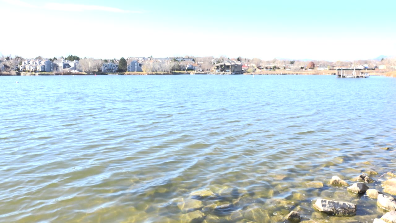 Opening view at Hidden Lake, Colorado with bright rippled water, rocks at the lower right, distant shoreline buildings, and no visible people.
