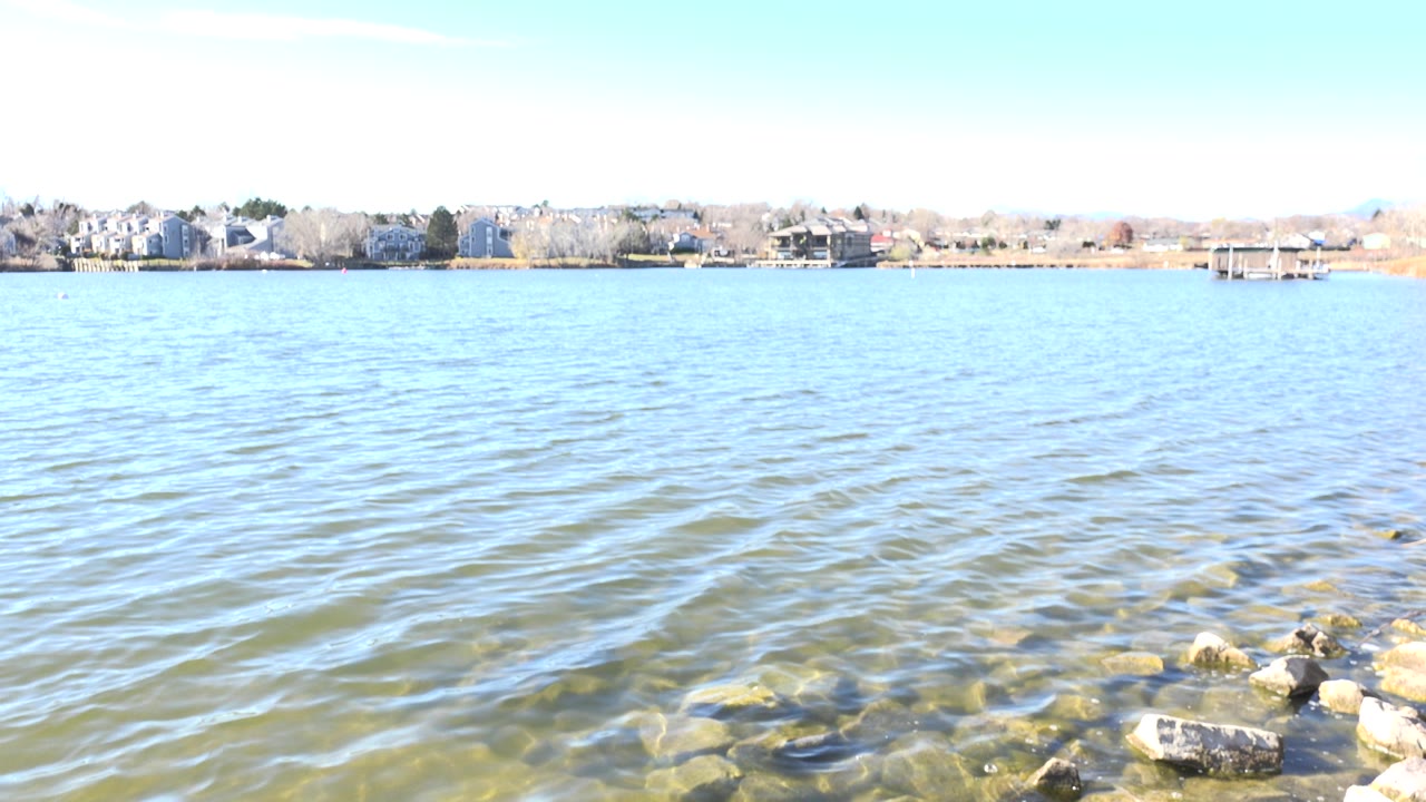 One second into the Hidden Lake, Colorado clip, small ripples cross the blue-green water while shoreline rocks and far buildings remain in view, with no visible people.
