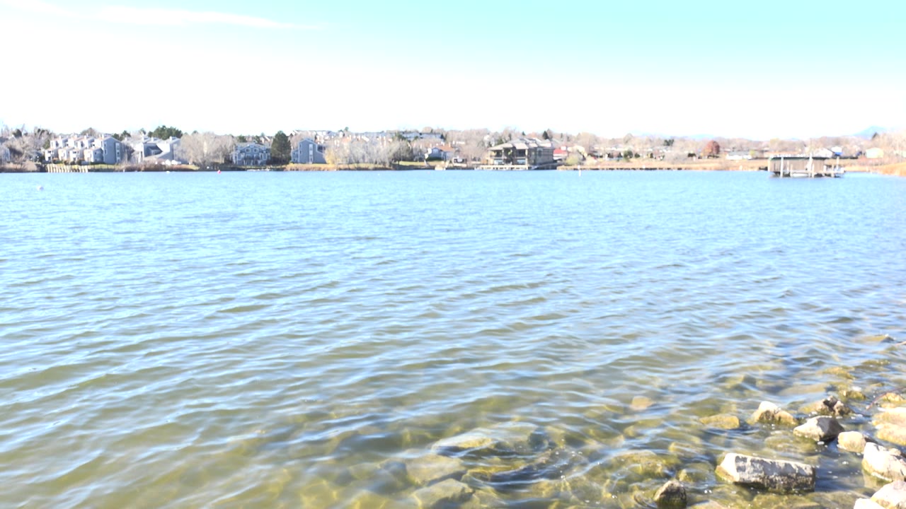 Second frame of Hidden Lake, Colorado showing small ripples across the water, a cluster of foreground rocks, distant residential buildings, and no visible people.