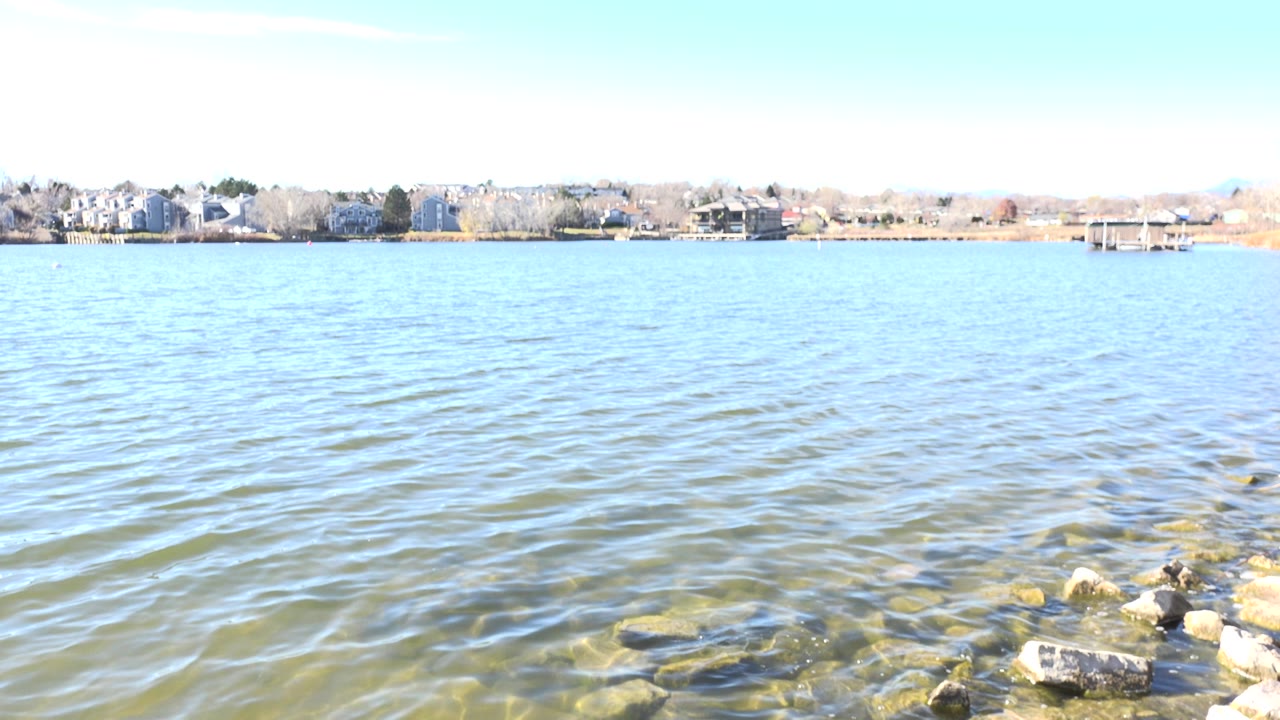 Lake water and rocky shoreline with distant homes under bright sky