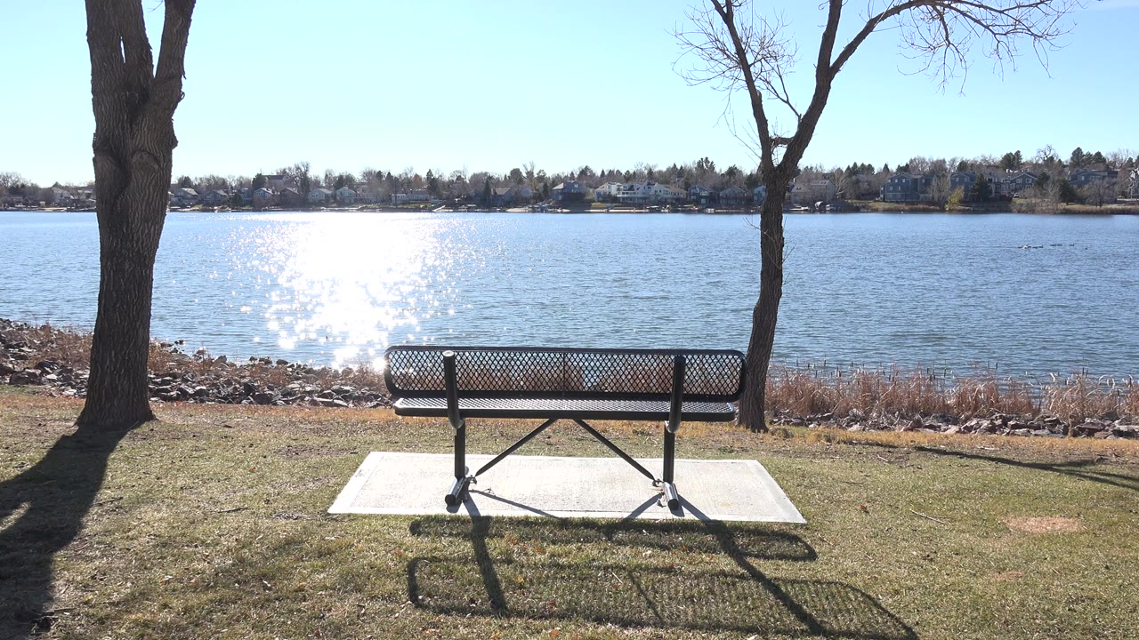 Opening frame of an empty bench facing Hidden Lake in Colorado, with bright sun glare on the water, bare trees at both sides, distant homes across the shore, and no visible people.