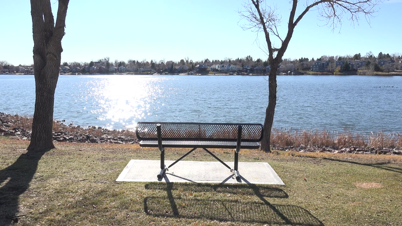 Opening frame at Hidden Lake, Colorado with a bench on a concrete pad facing bright lake water, tree trunks on both sides, distant shoreline buildings, and no visible people.